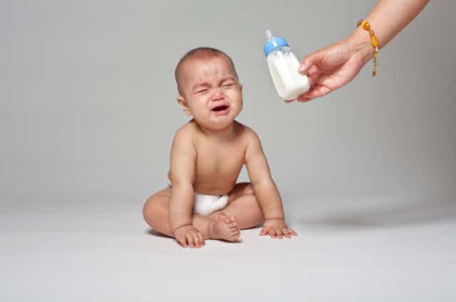 Baby in diaper sits on floor crying while a hand offers milk