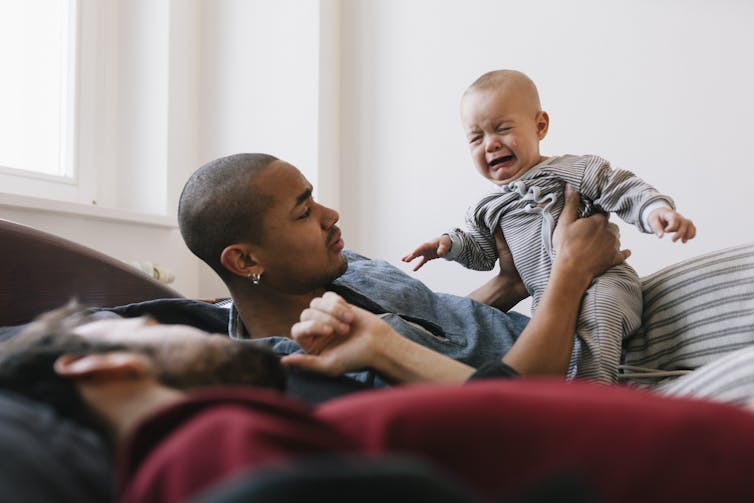 Gay fathers trying to calm a crying newborn. The group is lying on bed with large window and sunlight in the background.