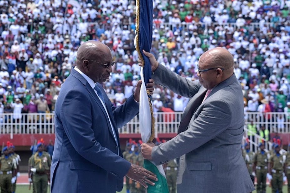A man hands over a folded national flag of Lesotho to another man at a crowded stadium.