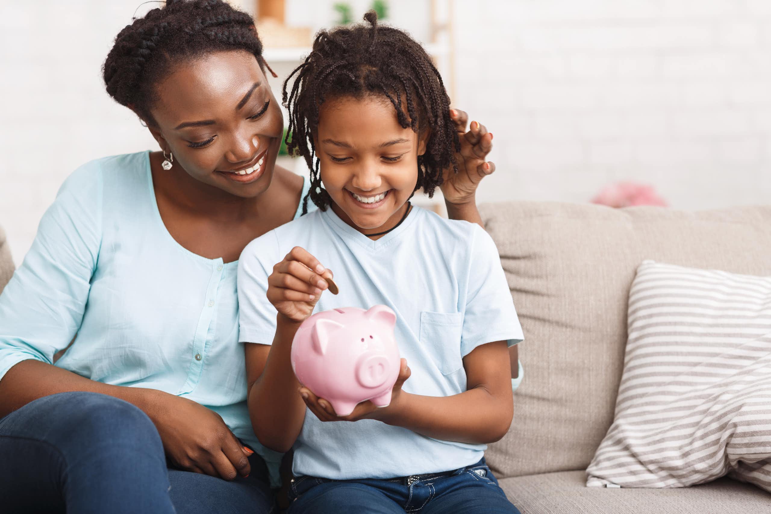 A woman in a light blue shirt and jeans smiles as her child, dressed in a similar outfit, puts money into a piggybank