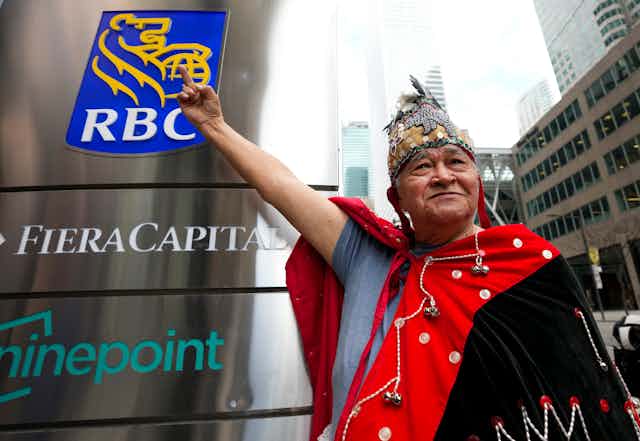 A man in Indigenous garb raises his middle finger to an RBC sign