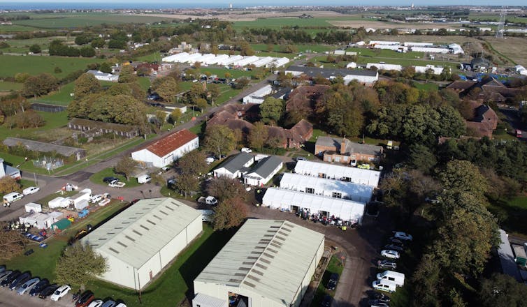 White tents and buildings among fields with the sea on the horizon.