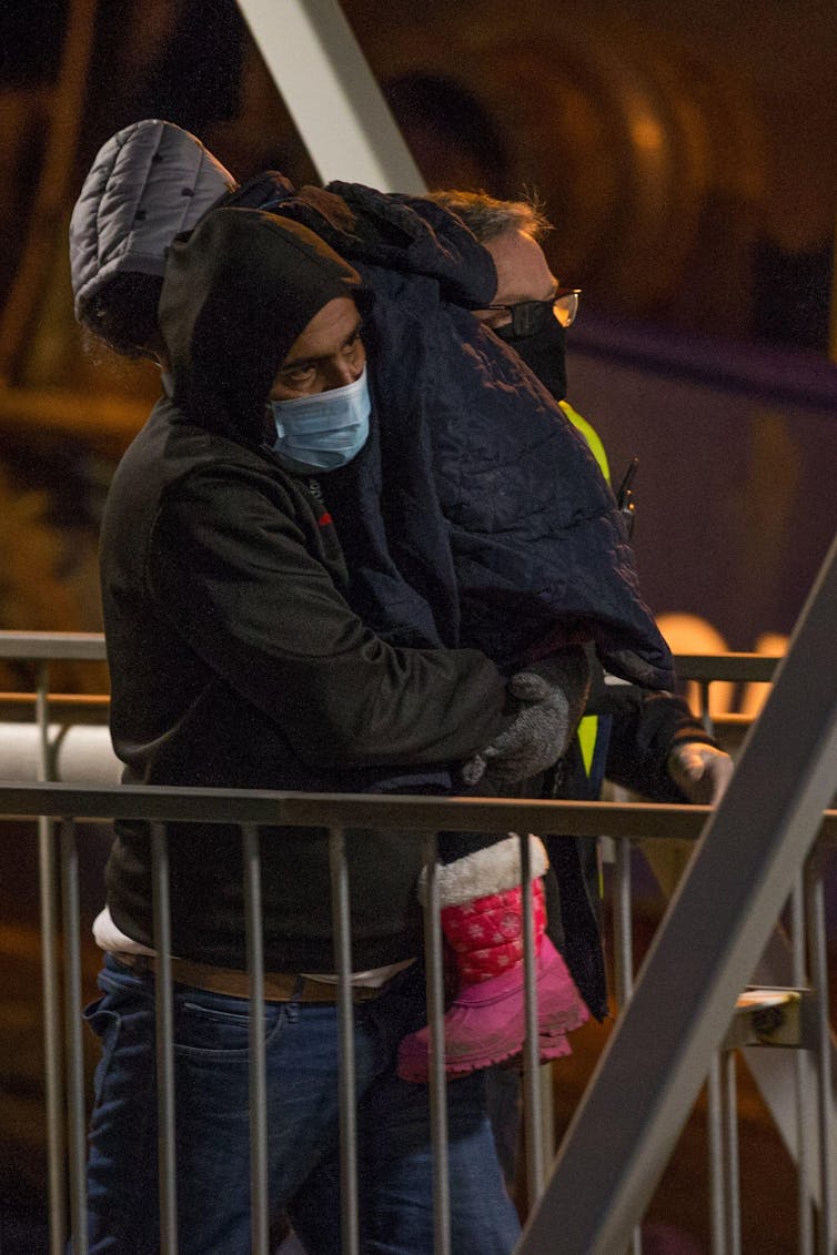 A man in a facemask carries a child wearing pink wellies and a hooded coat.