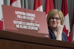A woman with short blond hair and glasses sits behind a placard that reads 'supporting canadians for a safe restart' in english and french