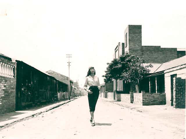 A sepia toned photograph of a glamorous young woman with long hair in a pencil skirt and blouse posing in the middle of a dirt street, gazing into the distance, her feet crossed