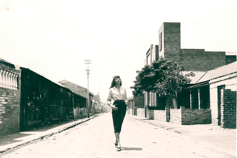 A sepia toned photograph of a glamorous young woman with long hair in a pencil skirt and blouse posing in the middle of a dirt street, gazing into the distance, her feet crossed