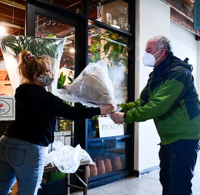 A florist hands a wrapped bouquet of flowers to a customer. Both wear face masks.