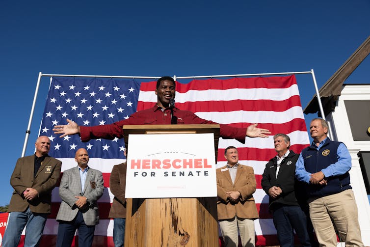 A Black man stands in a red shirt with his arms outstretched on a stage with several white men around him. He speaks at a podium with a banner that says Herschel for Senate.