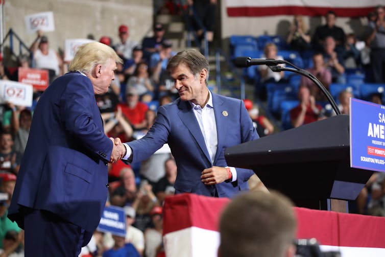 Two older men shake hands in blue suits, with a crowd of people behind them hold signs that say 'Oz' and 'Save America.