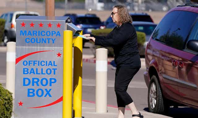 A woman in sunglasses drops something into a grey ballot drop box 