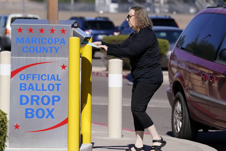 A woman in sunglasses drops something into a grey ballot drop box