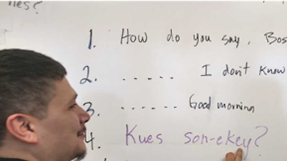 A teacher writes some words in an Indigenous language on a white board while a student looks.