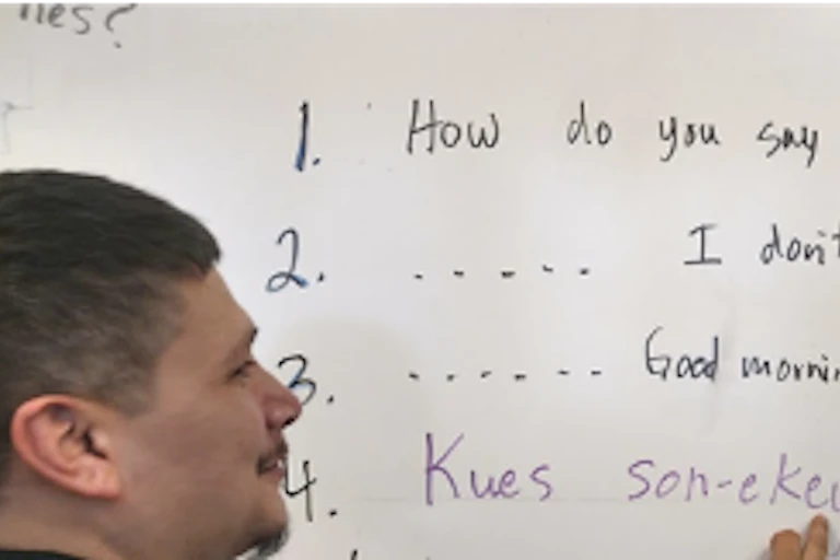 A teacher writes some words in an Indigenous language on a white board while a student looks.