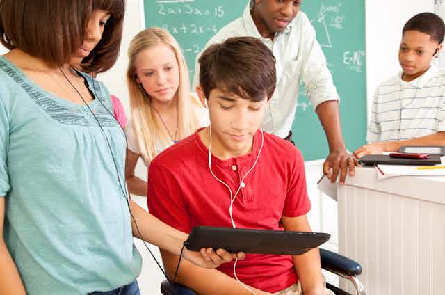 A student looks at an iPad held by another studenet while a teacher and other students stand in the background in front of a chalkboard with some math problems.