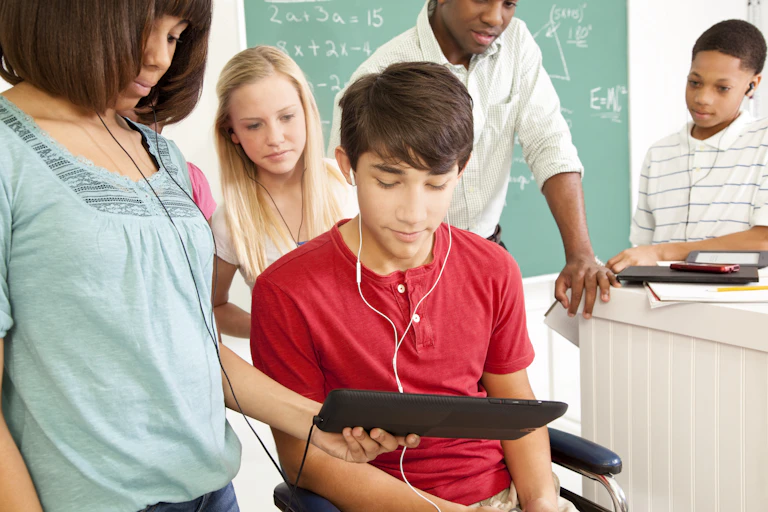 A student looks at an iPad held by another studenet while a teacher and other students stand in the background in front of a chalkboard with some math problems.
