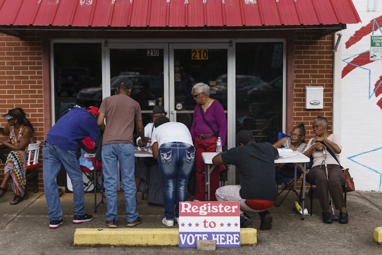 People standing at a table behind a sign that says'Register to vote here.'