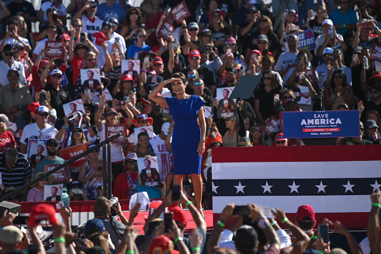 A white woman in a blue dress looks into the distance, with a large crowd of people behind her holding signs that say things like 'Save America'