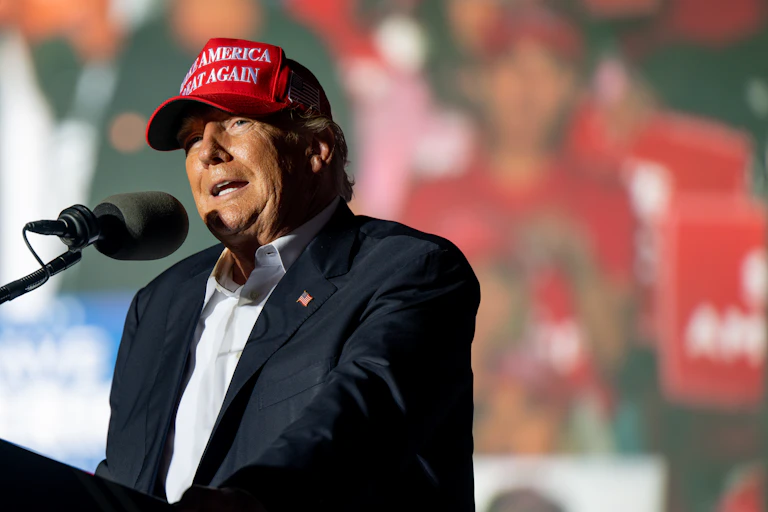 A man in a blazer and collared shirt wearing a red cap stands in front of a microphone