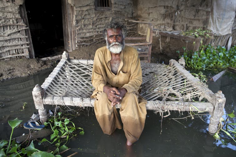 A man sits on a bench outside the door too his home, surrounded by floodwater up to his shins.