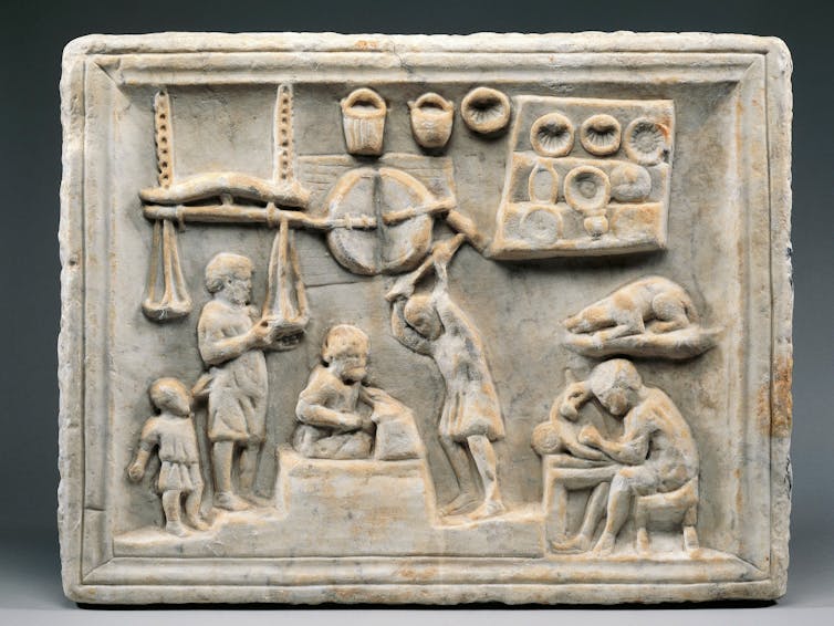 A carved scene shows a shop with bowls on the wall, a man hammering an object with a large hammer, another person writing, a dog and a child.