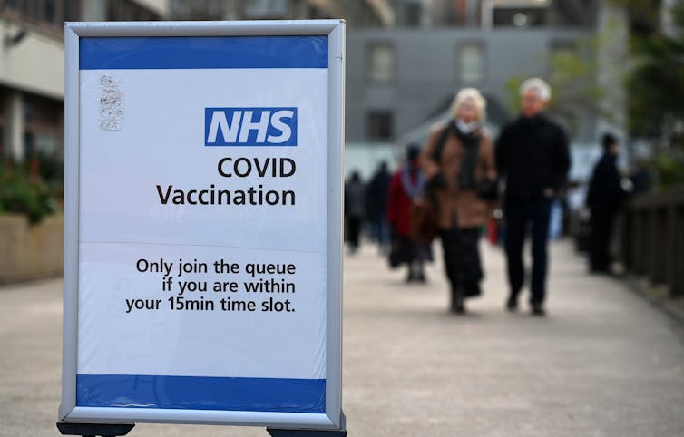 An NHS COVID vaccination sign in the foreground, people walking in the background.