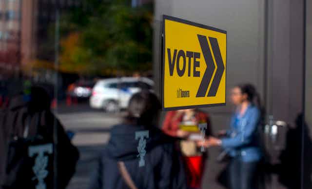 A yellow 'vote' sign is seen with people reflected in the glass behind it.