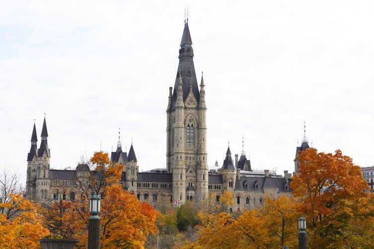 The West Block of Parliament Hill is pictured with orange, yellow and red trees framing it.