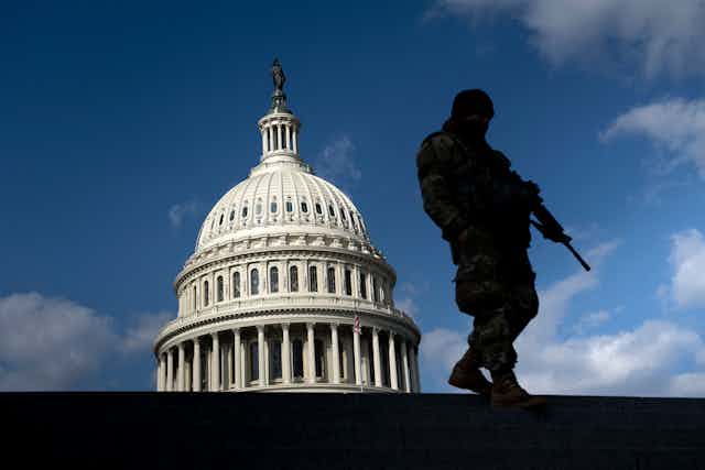 A man highlighted in black silhouette holds a gun and stands on a building across from the US Capitol, against a blue sky 