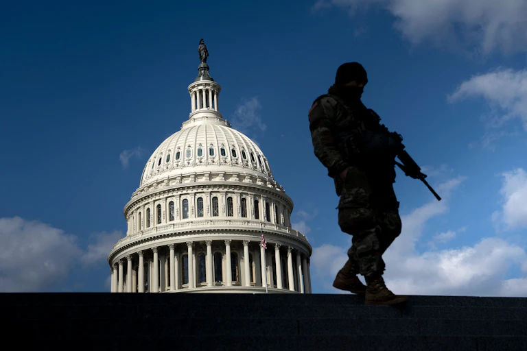 A man highlighted in black silhouette holds a gun and stands on a building across from the US Capitol, against a blue sky
