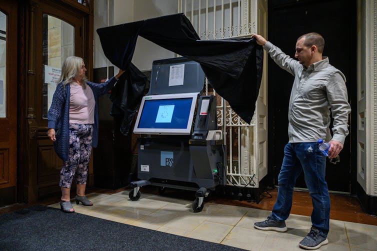 A white woman and man pull back a black curtain to show a voting machine with a big screen.