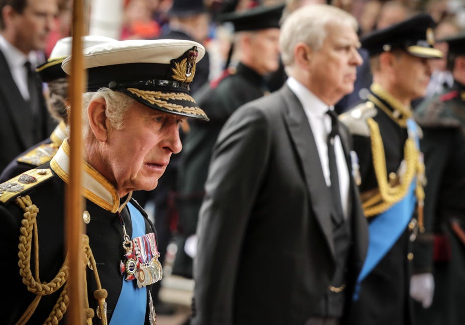 King Charles in military dress during the Queen's funeral procession, with Prince Andrew in a suit walking beside him, out of focus