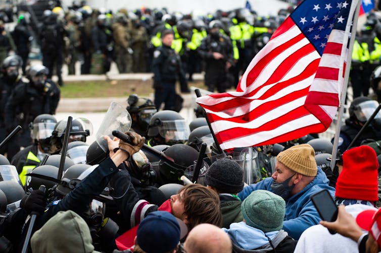 A crowd of people, some wearing protective helmets, push up against a group of protesters. One of them holds an American flag in the air.