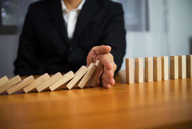 A person in a suit uses their hand to stop a domino pile of wooden blocks from falling
