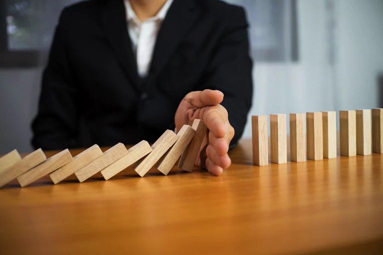 A person in a suit uses their hand to stop a domino pile of wooden blocks from falling