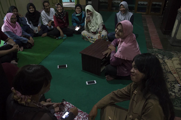 Women wearing colorful headscarves seated in a circle on a green carpet.