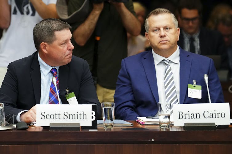 Two men in suits sit behind a conference table. The man on the left sits behind a name plate that says 'Scott Smith' and the man on the right sits behind a name place that says 'Brian Cairo'