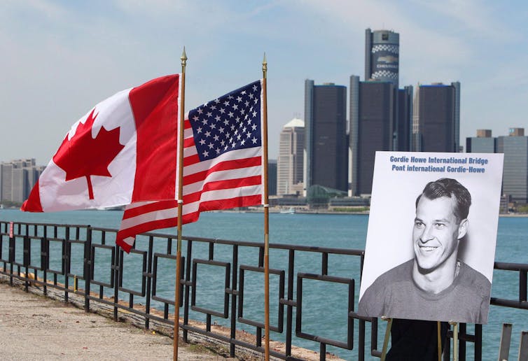 A black-and-white photo of a smiling man sitting on a bridge beside a Canadian flag and a U.S. flag