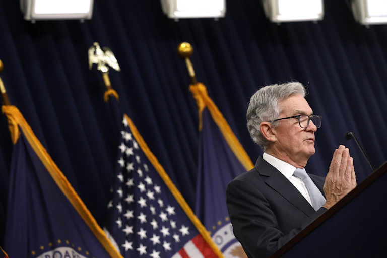 Fed chair Jerome Powell dressed in a grey tie and blue suit stand sin front of American flags.