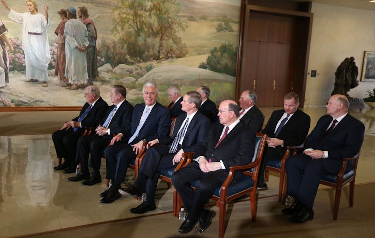 Two rows of men in suits sit in a formal room with a painting depicting Jesus in the background.