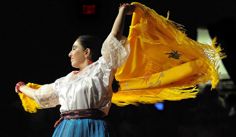 A brunette woman in a white top and blue skirt waves a yellow scarf as she dances.