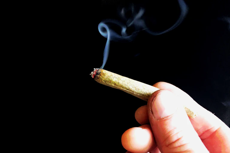 As a light trail of smoke wafts through the air, a hand holding a marijuana joint appears against a black background.