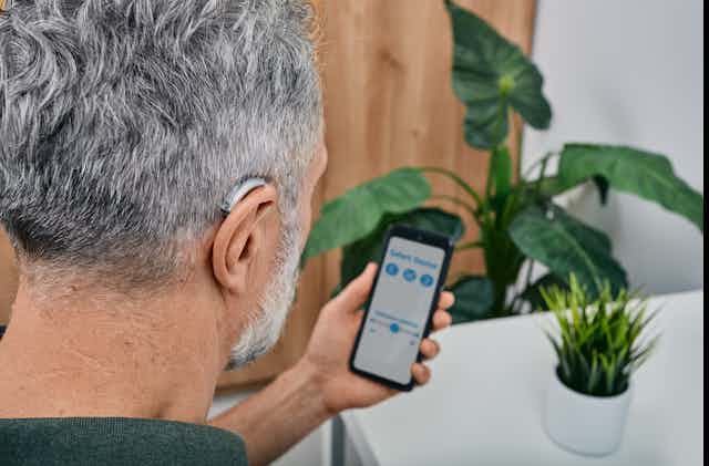 A hearing-impaired man with his back turned to the image adjusts settings for his hearing aid via the smartphone that he's holding.