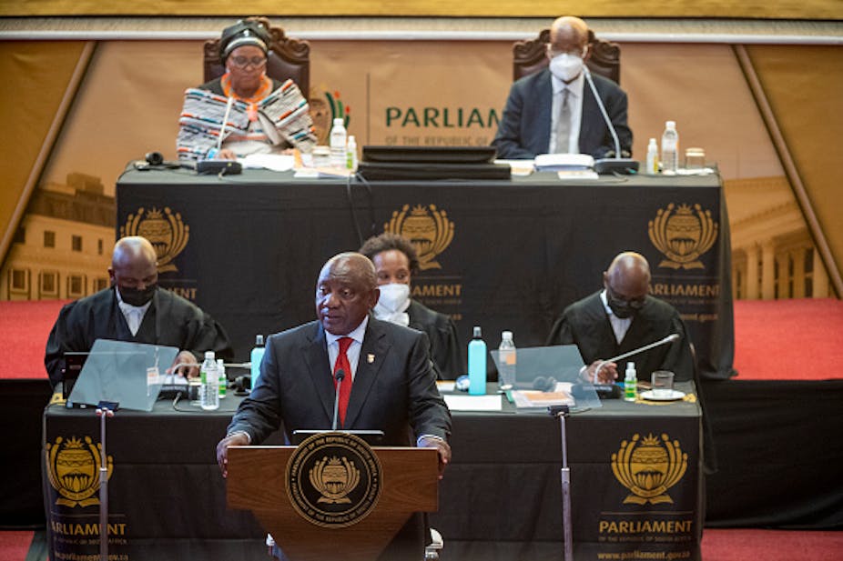 A man speaks with his hands balanced on a podium. Three man and two women sit behind him. 