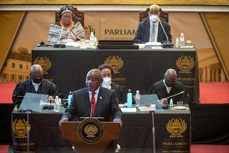 A man speaks with his hands balanced on a podium. Three man and two women sit behind him.
