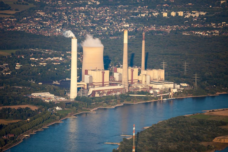 An aerial shot of a power station on the bank of a river, with a plume of smoke rising from the chimney.