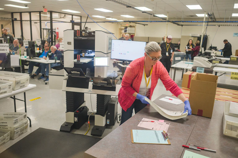 A person wearing gloves carries a stack of papers to a table, in the middle of a large room filled with tables and equipment