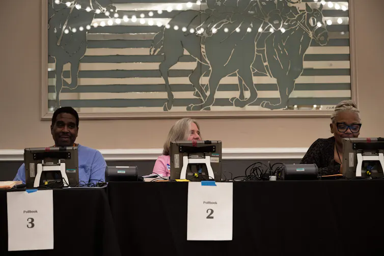 Three people sit behind a table, with computer screens in front of them.