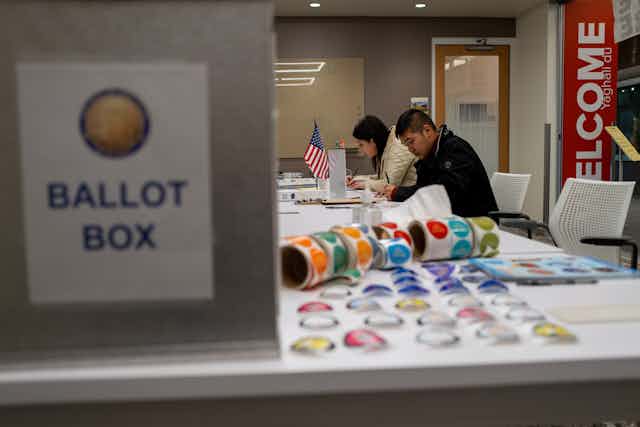 Two people are seated at a white table with stickers and an American flag, with a sign nearby that says ballot box.