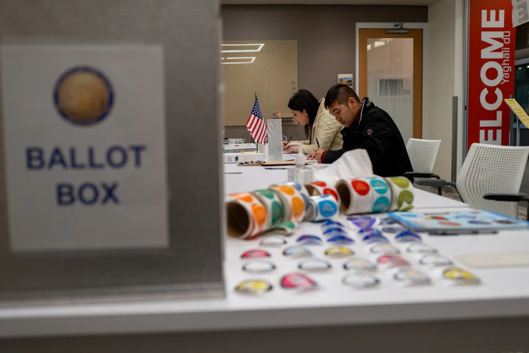 Two people are seated at a white table with stickers and an American flag, with a sign nearby that says ballot box.