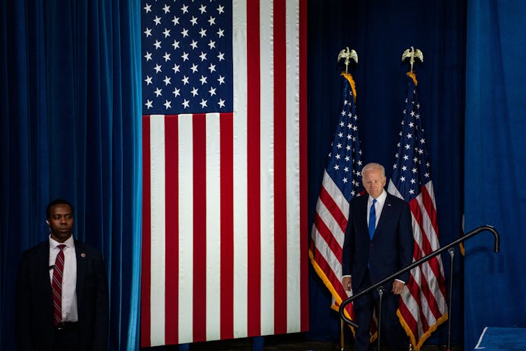 A white older man in a dark blue suit stands next to two American flags, and a third very large flag over a blue backdrop. A Black man in a suit stands on the other side of the American flag.
